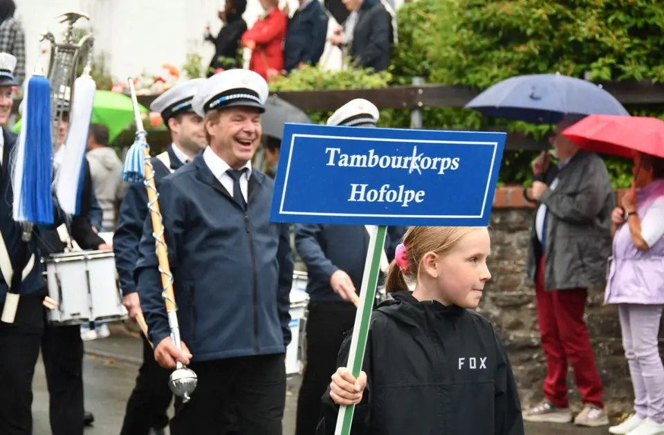 Ein Mädchen trägt bei Regen das Schild „Tambourkorps Hofolpe" an der Spitze des Zuges, dahinter der Tambourmajor mit Tambourstab und die Trommler
