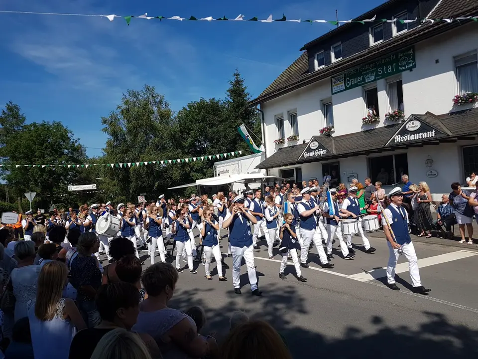 Das Tambourkorps zieht mit Flöten und Trommeln an den Zuschauern und dem Gasthaus Haus Höfer vorbei, über der Straße hängen grün-weiße Wimpelketten