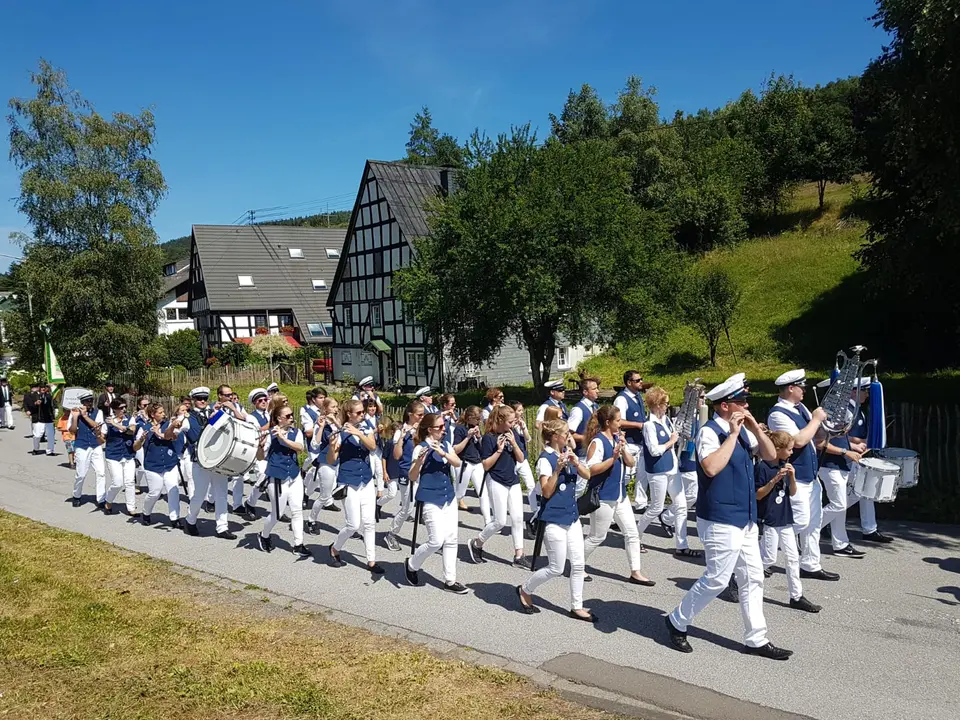 Das Tambourkorps Hofolpe marschiert bei Sonnenschein über eine Dorfstraße, im Hintergrund ein Fachwerkhaus und ein bewaldeter Hang